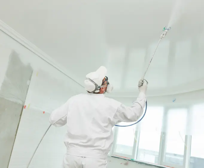 Painter in white coveralls spraying a ceiling in an empty room